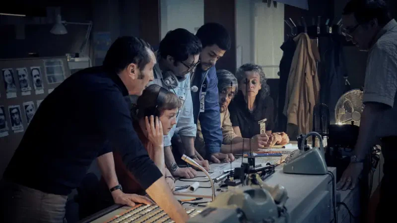 A group of filmmakers collaborates intensely around a table filled with equipment and notes, capturing a behind-the-scenes moment in the making of a movie on September 5.