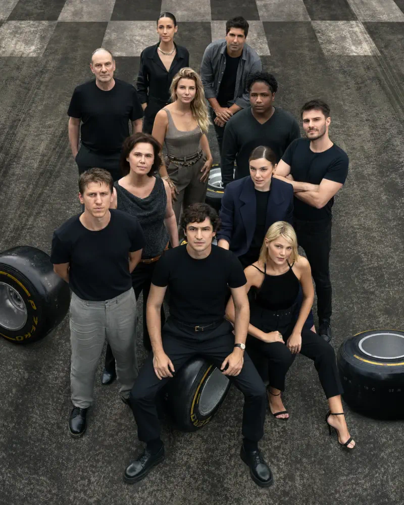 A cast of characters poses on a racetrack, surrounded by tires, in a promotional image for the TV show Senna (2024), showcasing a dynamic ensemble with varied expressions.