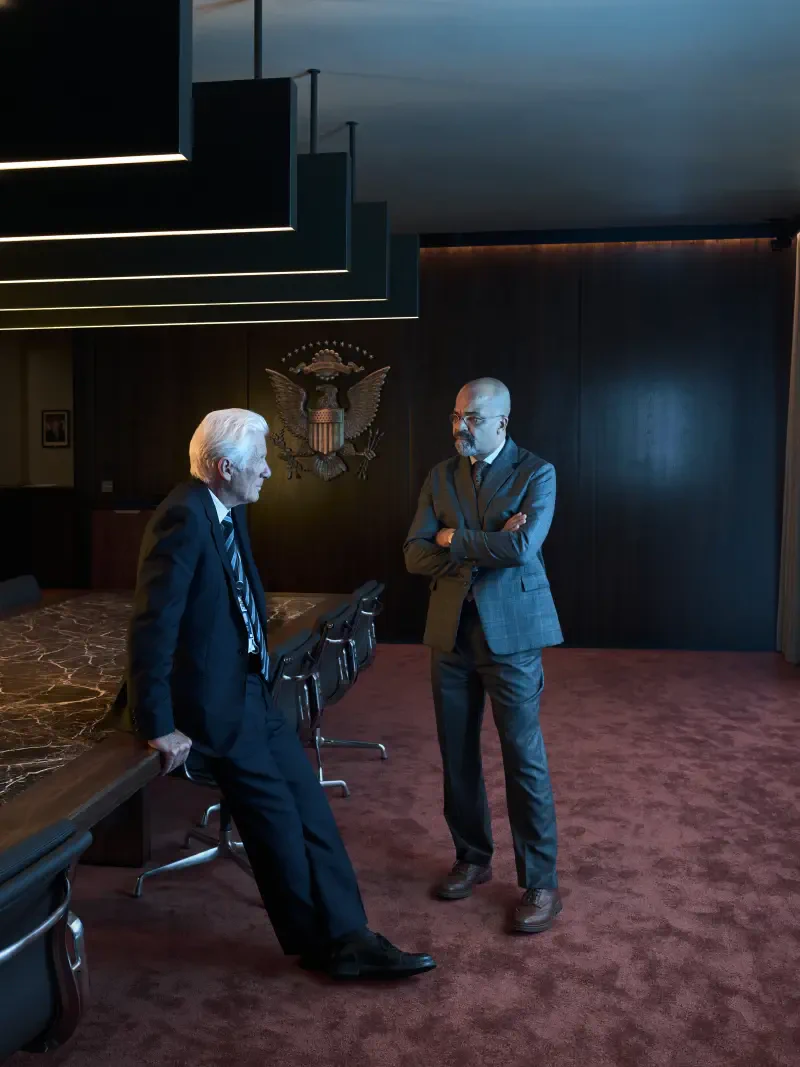 Richard Gere and Jeffrey Wright engage in conversation in a sleek, modern boardroom setting from the TV show The Agency. The ambiance is professional and tense.