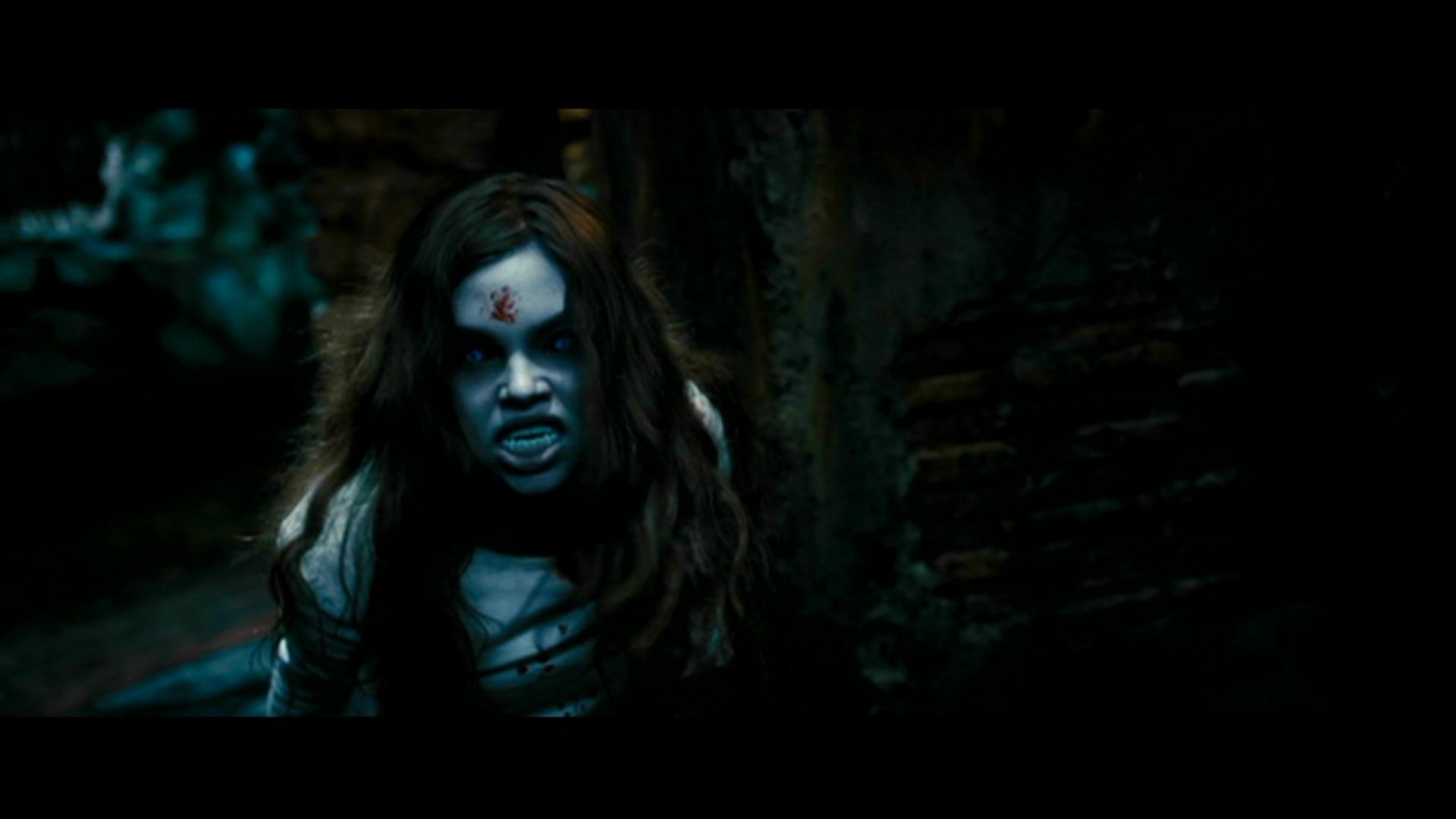 Blue-lit scene from the movie Underworld: Evolution showing a pale, bloodied woman with fangs and wild hair crouched against crumbling brick.