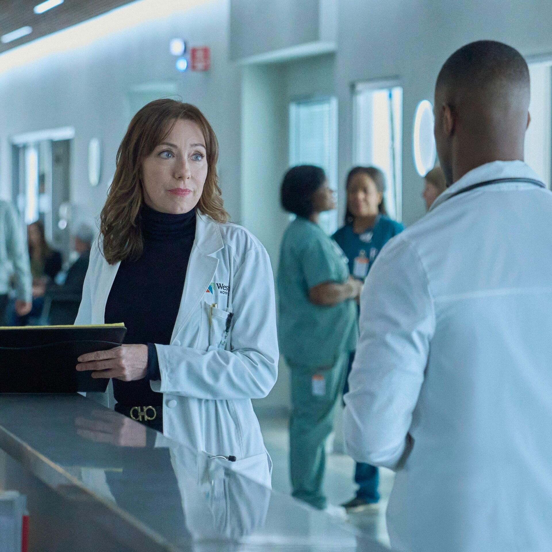 Molly Parker stars in the TV show Doc (2025), engaging with a colleague in a modern hospital setting, surrounded by staff in scrubs.