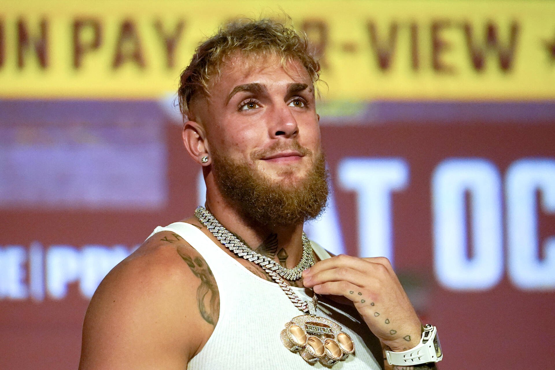 Jake Paul poses confidently in a boxing event, showcasing his distinctive style with a prominent beard and eye-catching jewelry against a promotional backdrop.