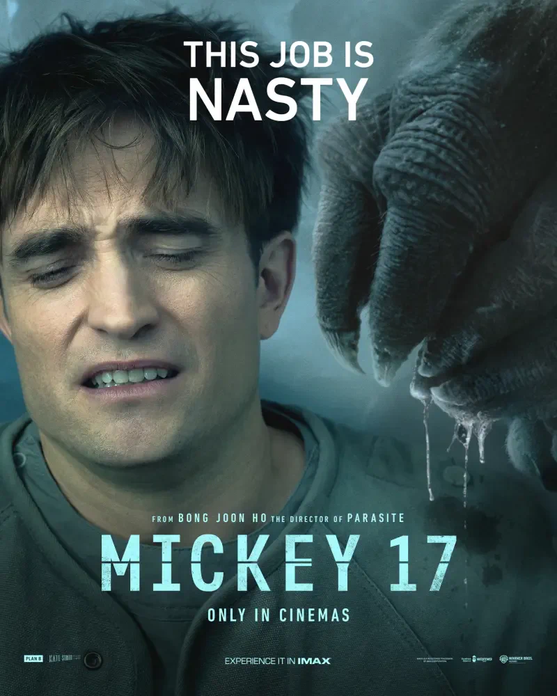 A still from the film Mickey 17 (2025) featuring Robert Pattinson with an expression of discomfort, as a large hand looms ominously above him, emphasizing the film's tense atmosphere.