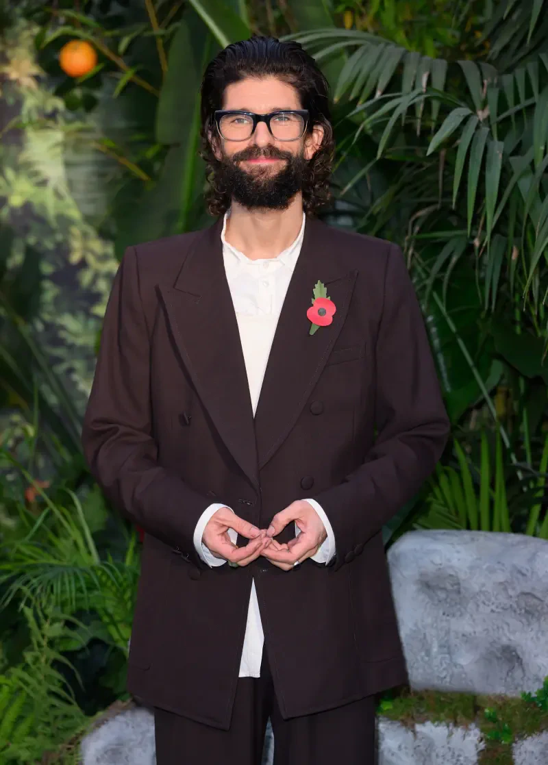 Ben Whishaw poses in a stylish brown suit, celebrating the movie Paddington in Peru, surrounded by lush greenery and tropical elements.