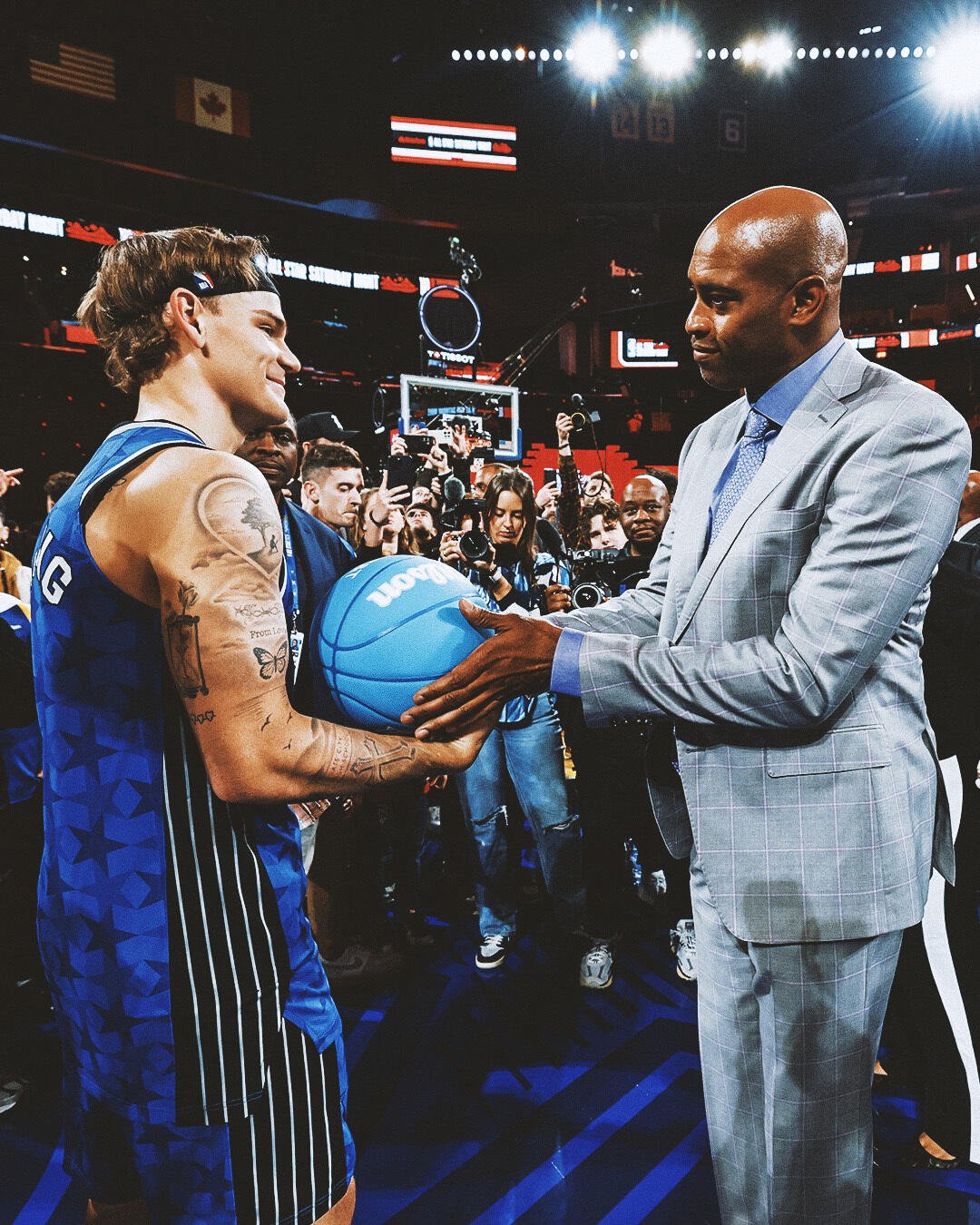 Mac McClung hands a basketball to Vince Carter amid a crowd at the NBA All-Star Game, highlighting the excitement of basketball legends and rising stars.