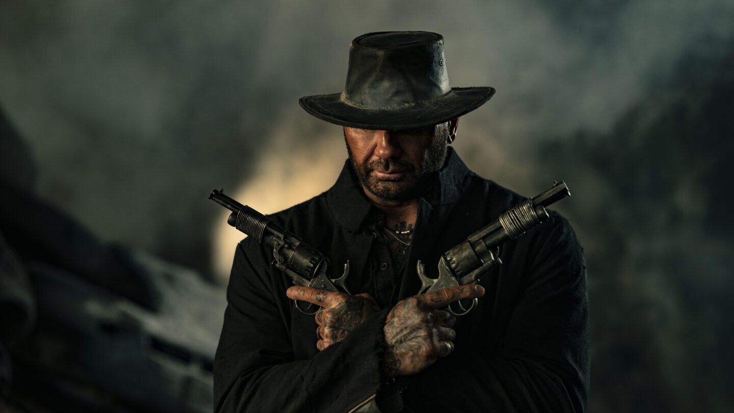 A gritty scene from the movie In The Lost Lands, featuring Dave Bautista in a dark outfit, holding two guns and wearing a wide-brimmed hat, set against a smoky backdrop.