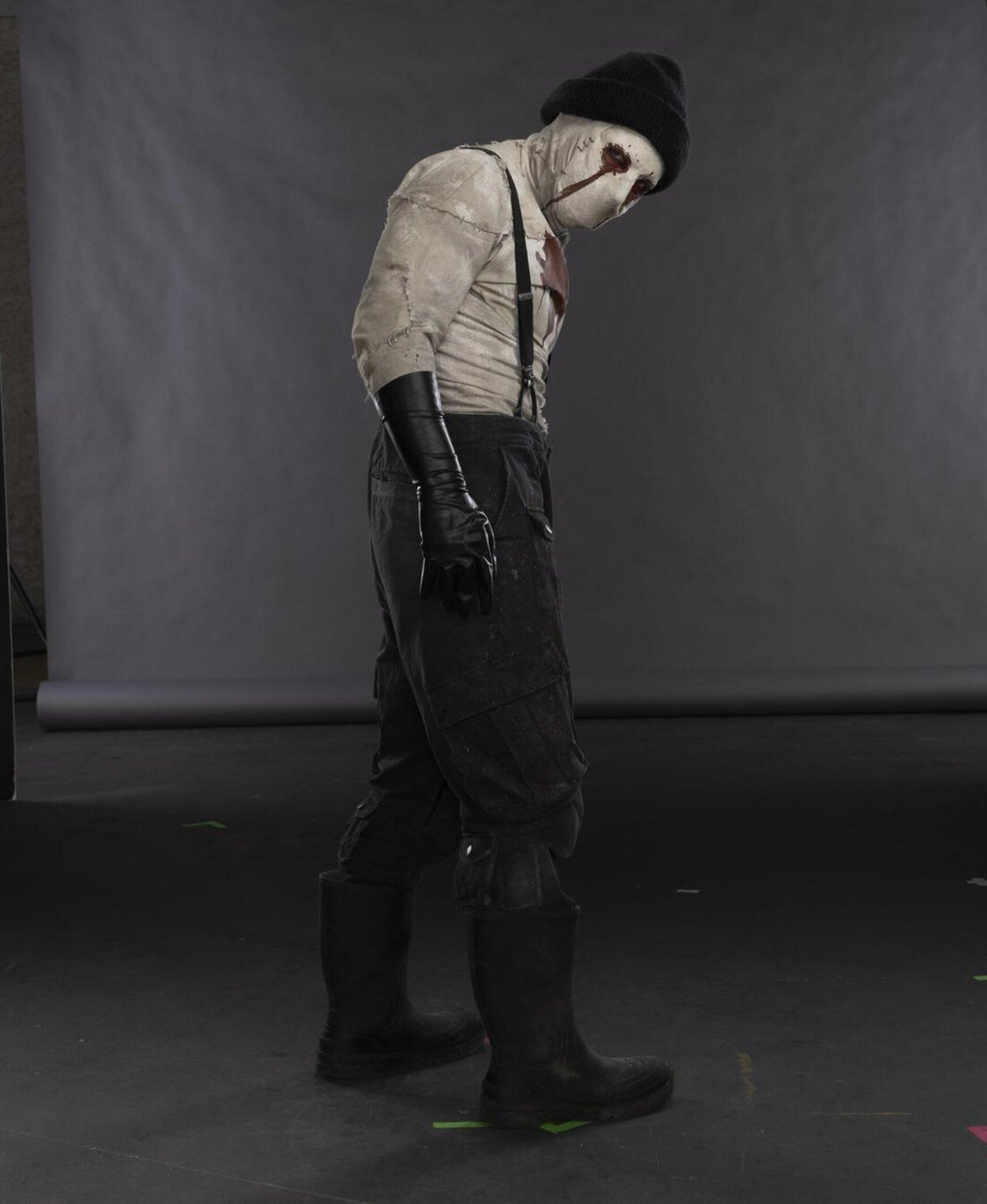 Bandaged, masked figure in suspenders, black gloves and boots walks across a gray studio set — promotional image for the TV show Daredevil: Born Again.