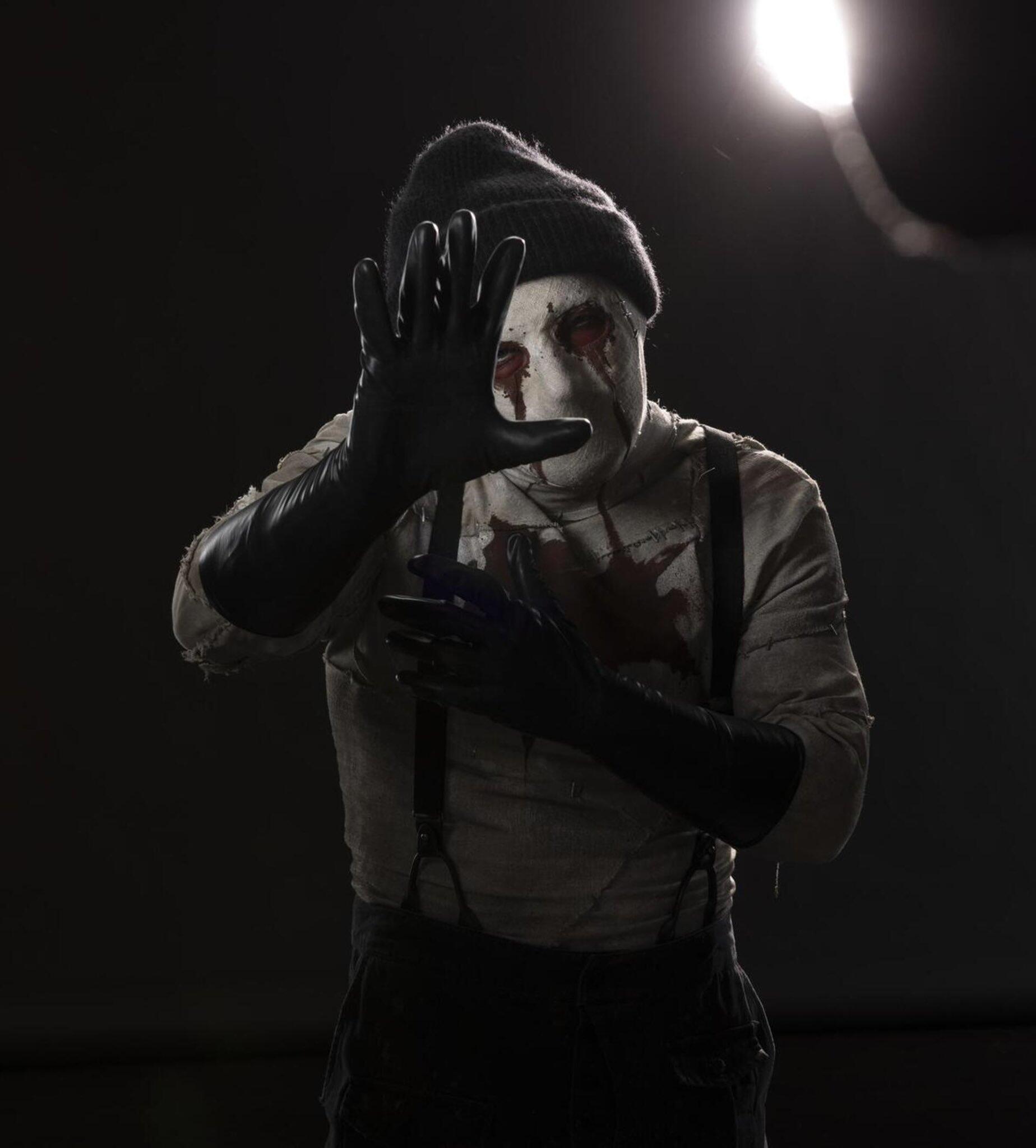 Promotional still from Daredevil: Born Again (TV show) — masked, blood-streaked figure in a beanie and gloves reaches toward the camera under a harsh spotlight.