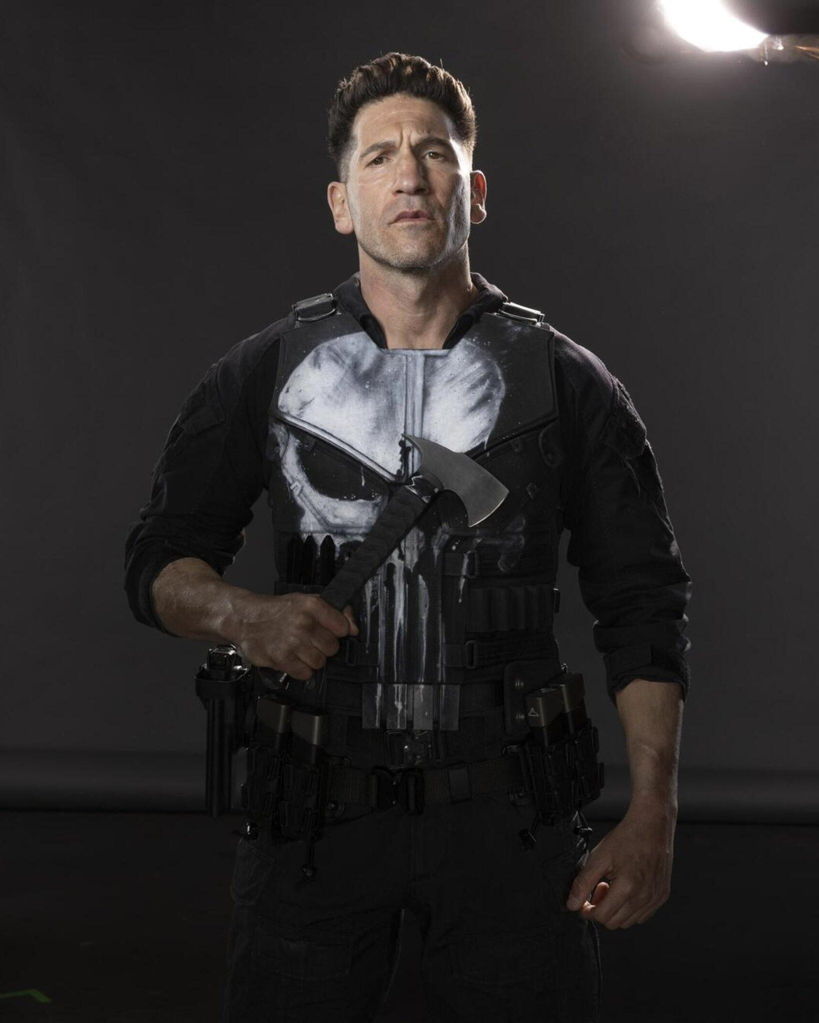Man in tactical vest with white skull emblem holding an axe, posed against a gray backdrop — promotional image for the TV show Daredevil: Born Again.