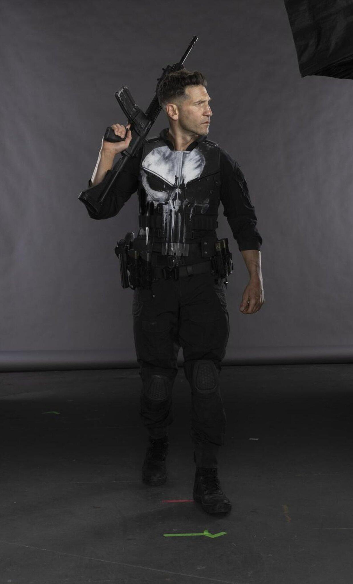 Figure in tactical black gear with a white Punisher skull and rifle walking on a studio set for Daredevil: Born Again (TV show).