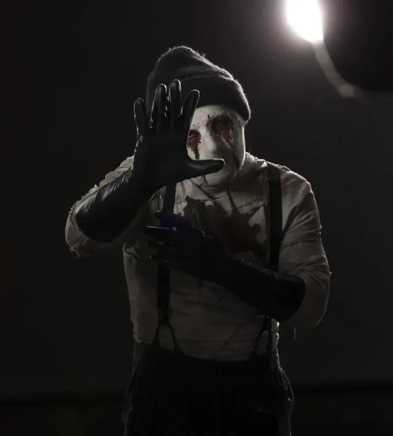 Promotional still from Daredevil: Born Again (TV show) — masked, blood-streaked figure in a beanie and gloves reaches toward the camera under a harsh spotlight.
