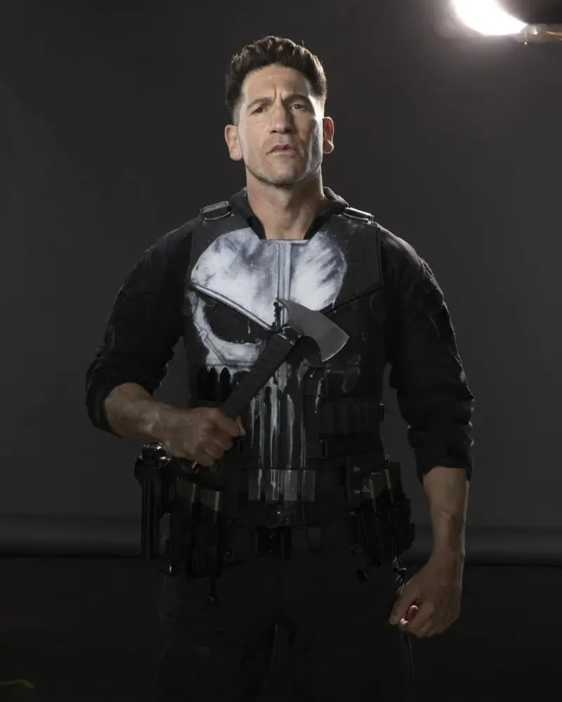 Man in tactical vest with white skull emblem holding an axe, posed against a gray backdrop — promotional image for the TV show Daredevil: Born Again.