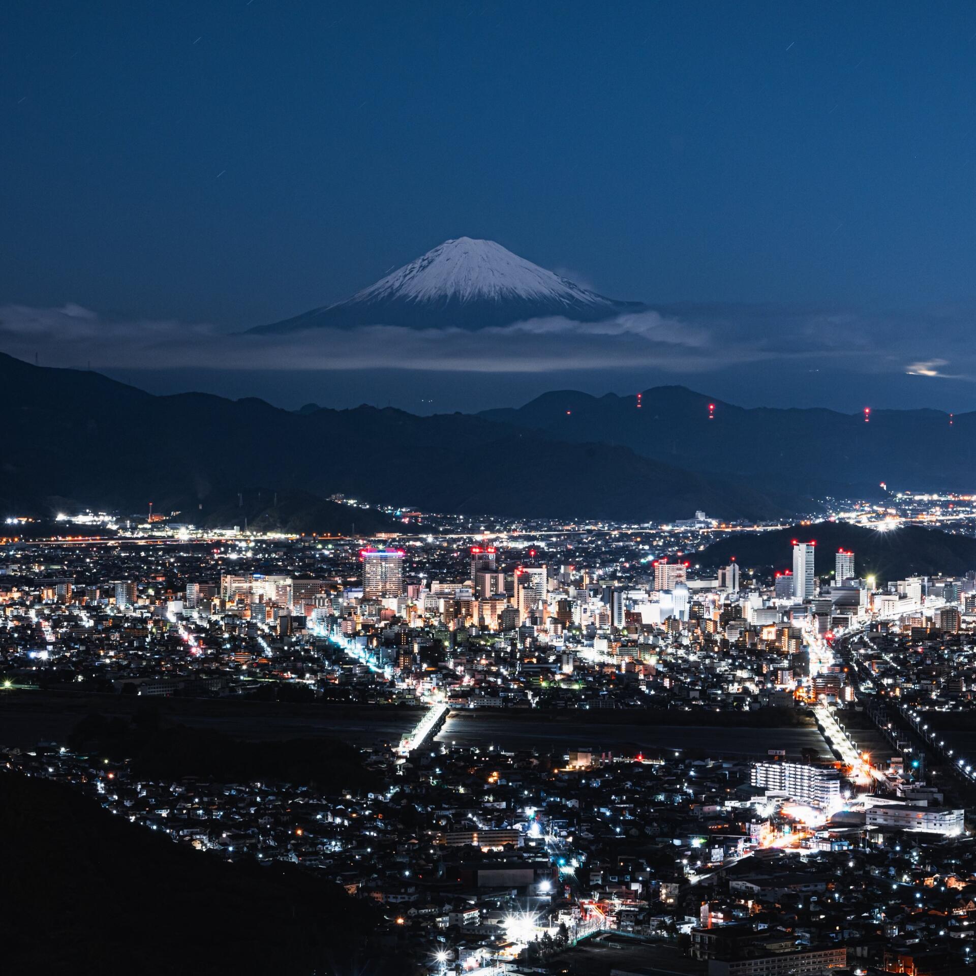 Night cityscape of Yamanashi, Japan, glowing streets and buildings with snow-capped Mount Fuji looming above a low band of clouds.