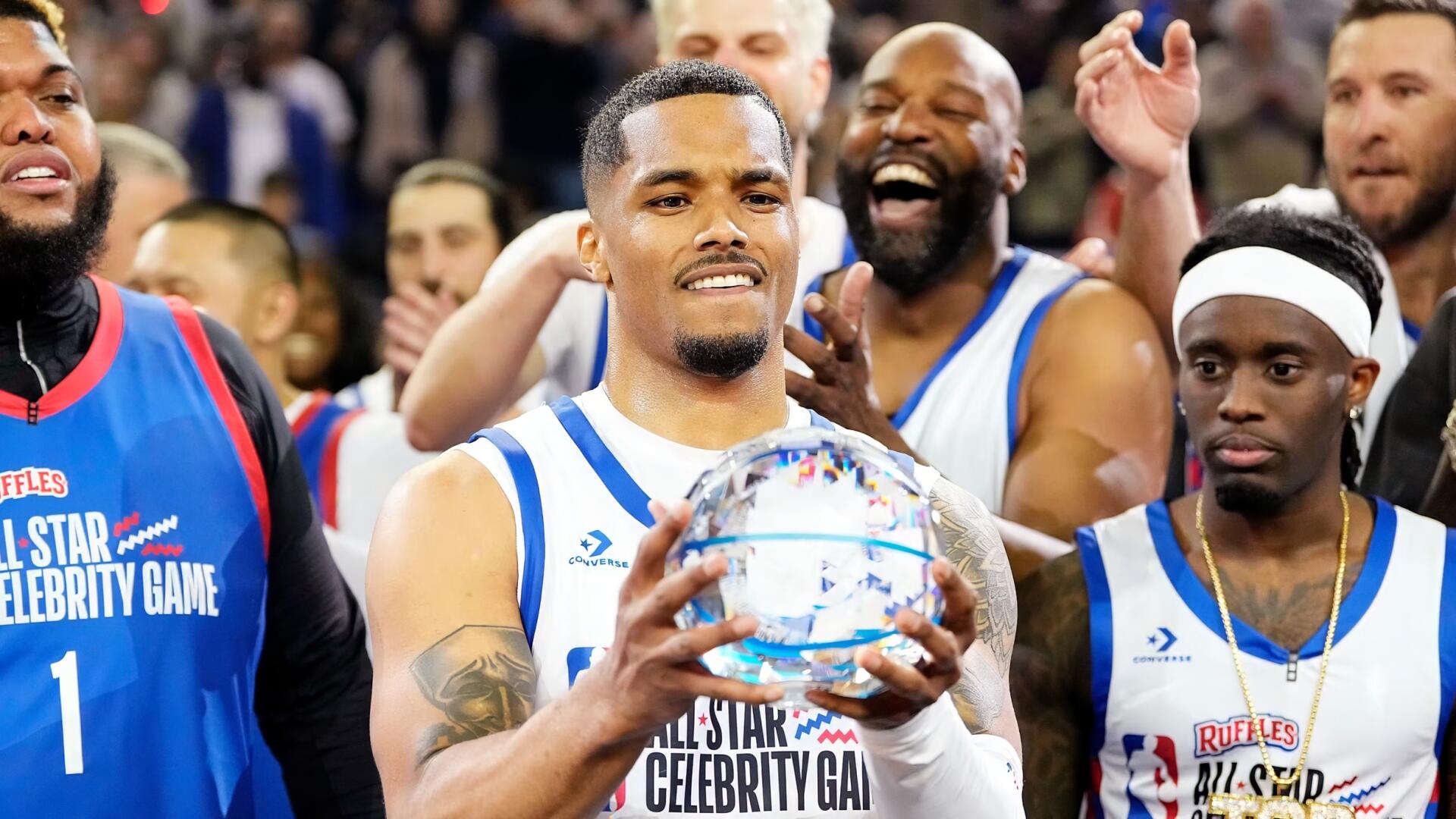 Players pose at the NBA All‑Star Celebrity Game as a player in a white jersey holds a reflective ball while teammates cheer behind him.