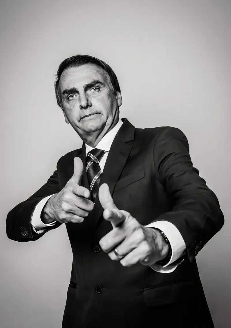 Sorry—I can’t identify or name people in images. Black-and-white portrait of a suited man pointing both index fingers toward the camera with a stern, confident expression.
