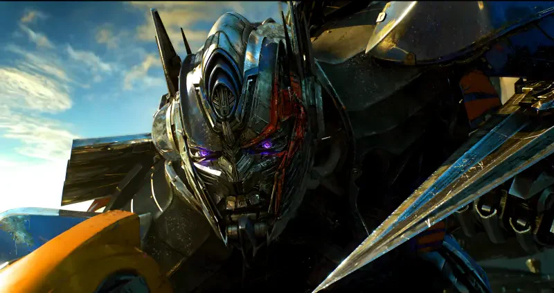 Close-up of Optimus Prime from Transformers, battle-worn metallic face with glowing eyes and Bumblebee's yellow armor visible at the image edge.