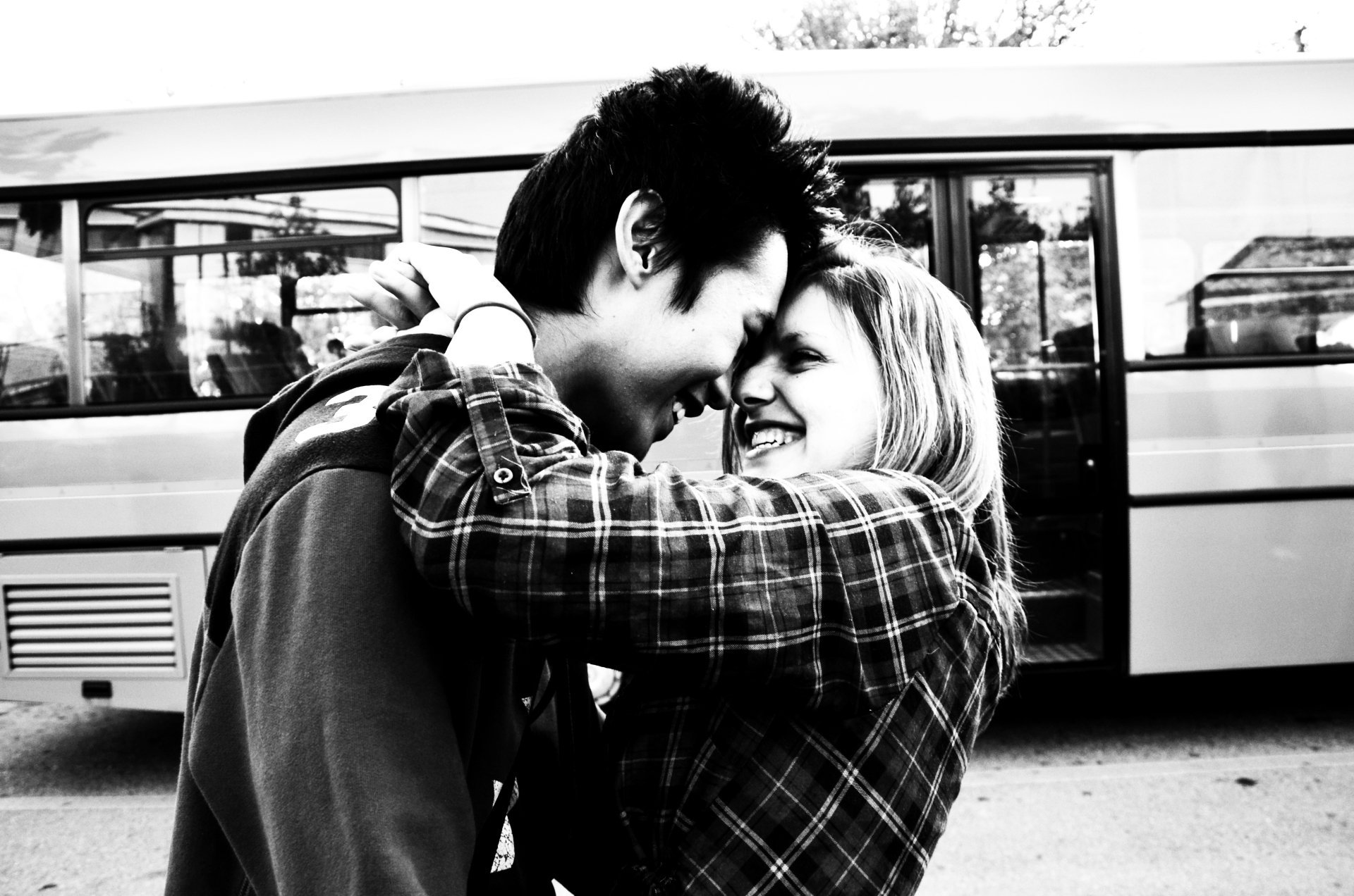 A black-and-white photograph captures a joyful moment between a young couple embracing and laughing, with a bus in the background, showcasing a candid, intimate atmosphere.