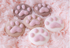 A close-up of decorative cookies shaped like animal paws, featuring shades of pink and brown, arranged on a fluffy pink background.